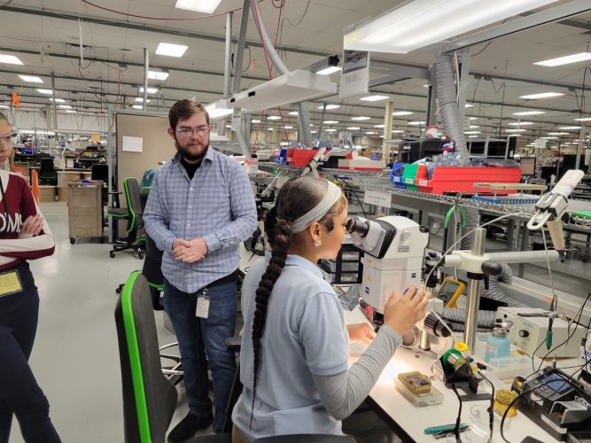 Student looking at the detailed soldering on a component for a video borescope at Waygate Technologies, accompanied by James Mark (left), a graduate of the Auburn P-TECH program. 